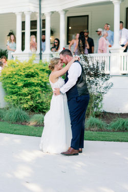 Bride and groom embracing on the lawn in front of a white porch as guests look on